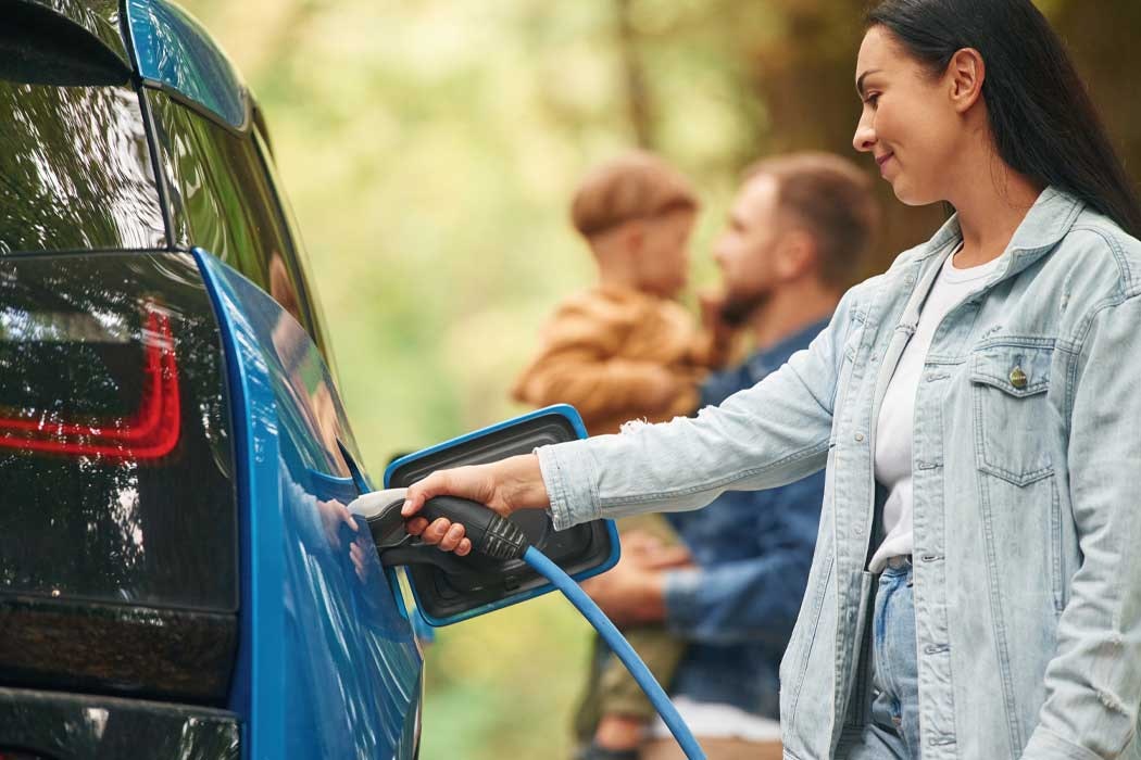 Auf dem Bild lädt eine junge Frau mit dunklen, langen Haaren ein Elektroauto auf. Sie trägt eine hellblaue Jeansjacke über einem weißen T-Shirt. Mit ihrer rechten Hand hält sie das Ladekabel, das in die Ladestation des Autos gesteckt ist. Sie steht seitlich zum Auto, schaut aufmerksam auf das Ladegerät und scheint das Laden zu überwachen. Im Hintergrund sind zwei Männer zu sehen, die ein Kind halten. Die Männer haben kurze Haare und sind im Gespräch miteinander vertieft. Der Hintergrund zeigt eine natürliche Umgebung mit Grünflächen und Bäumen, was auf einen Park oder Wald hinweisen könnte.
