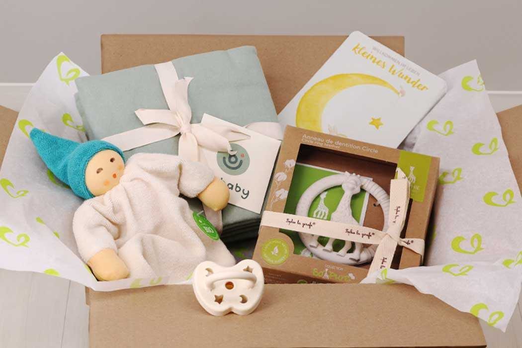 Das Bild zeigt einen offenen Karton, gefüllt mit Babygeschenken. Im Inneren liegt ein kleines, weiches Stofftier, das ein Baby mit blondem Haar, in einem weißen Schlafanzug und mit einem blauen Mützchen darstellt. Neben dem Stofftier befindet sich eine hellblaue Wickelunterlage mit weißer Schleife, sowie verschiedene Babyartikel in einer Verpackung. Darunter sind ein weiches, weißes Lätzchen mit einem Sternmuster und eine Baby-Halskette oder -Schnullerkette, die in einer braunen Verpackung mit transparentem Fenster steckt. Der Karton ist liebevoll mit weißen Papierstreifen ausgelegt, die mit grünen Herzen bedruckt sind.