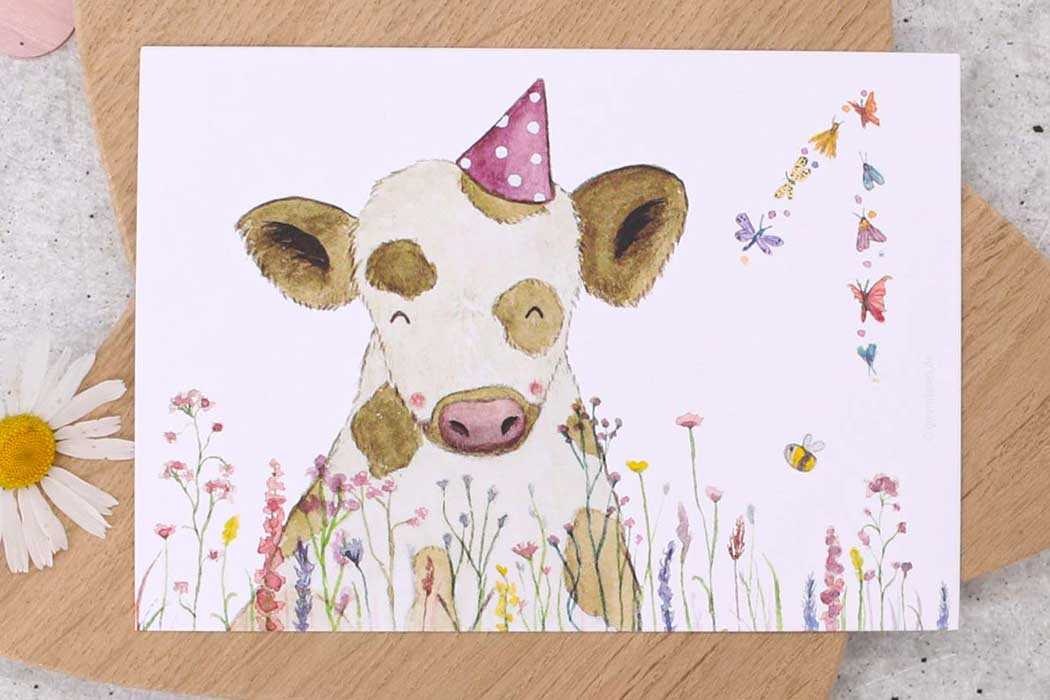 Das Bild zeigt eine fröhliche Geburtstagskarte, die auf dem Boden im Freien liegt, umgeben von grünen Pflanzen und Gras. Die Karte verfügt über eine liebevolle Illustration eines kleinen, süßen Schäfchens mit einem lachenden Gesicht und rosigen Wangen. Das Schäfchen trägt eine rosa Partyhütchen mit weißen Punkten. In der Umgebung des Schäfchens sind kleine, zarte Pflanzen und Blumen sowie zwei summende Bienen zu erkennen. Oben rechts auf der Karte steht in einem freundlichen, gelben Schriftzug: „Alles Liebe zum Geburtstag“, wobei das Wort „Geburtstag“ hervorgehoben ist.